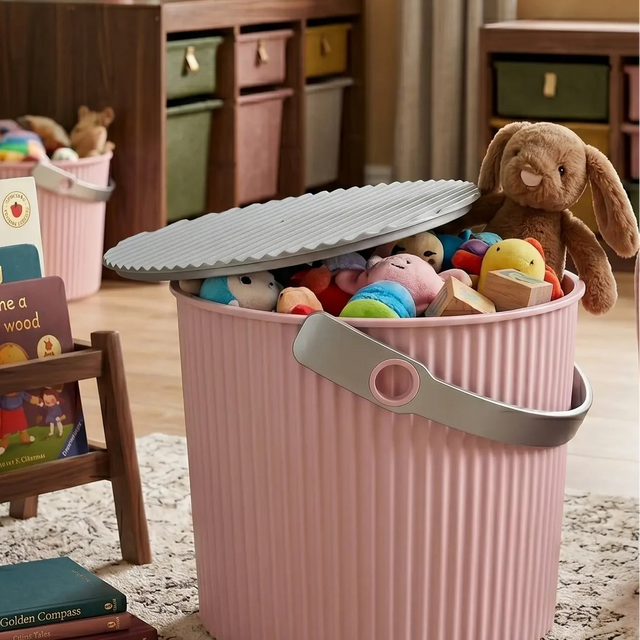 Rosa Aufbewahrungseimer aus Kunststoff mit Deckel im edlen Kinderzimmer im Skandi-Design. Der kleine Spielzeug-Eimer mit grauem Henkel dient als dekorative Spielzeugkiste für Kuscheltiere neben einem Sessel und einem Kinderbücherregal aus Holz.